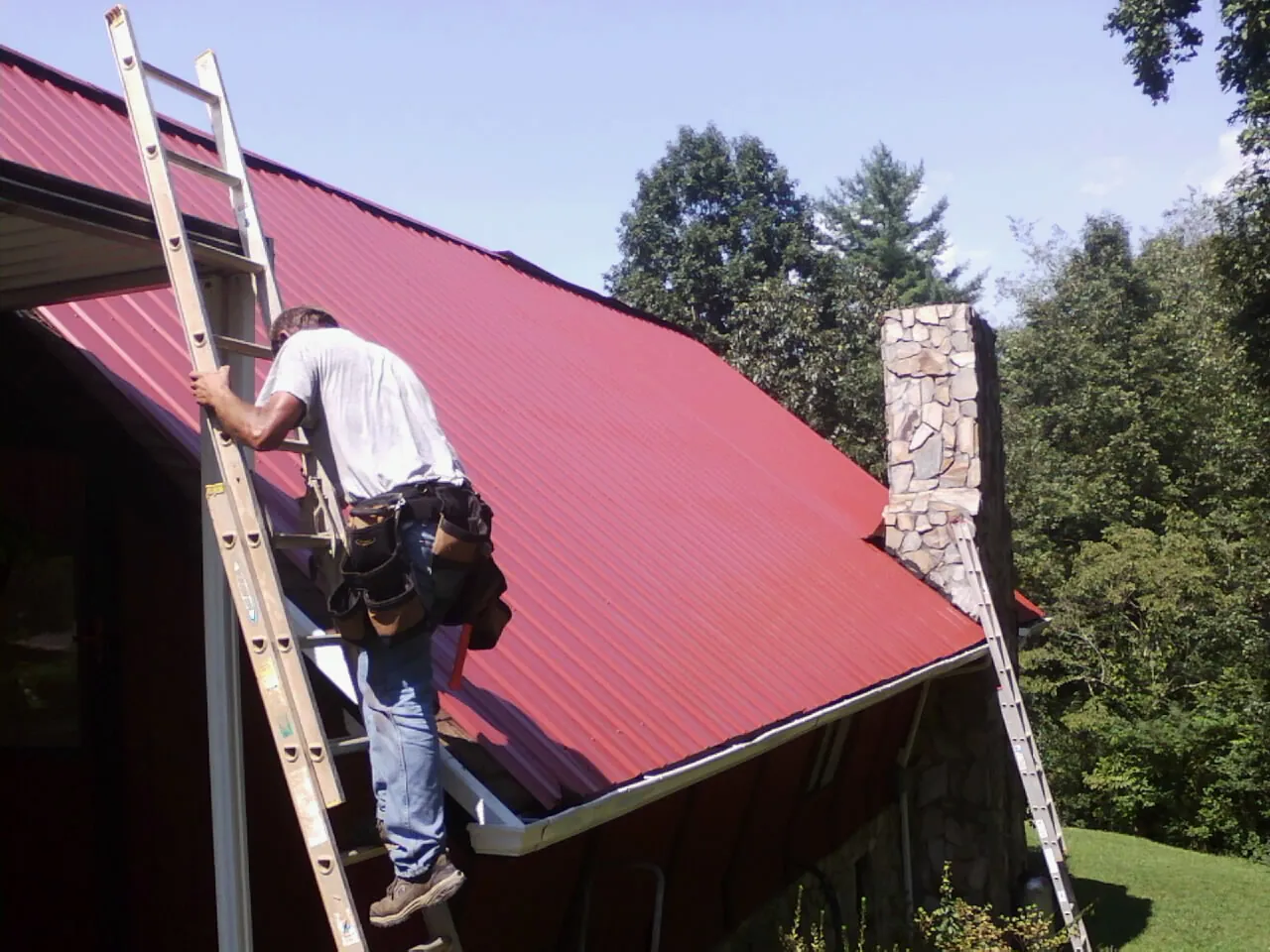 Roofer on a ladder beside a red metal roof and stone chimney — Soffit & Fascia Repair by Roofing Brothers in Kutztown